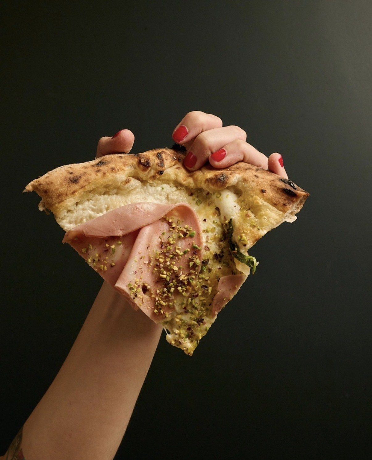 Fetta di pizza bianca con mortadella e granella di pistacchio tenuta in mano su fondo scuro
